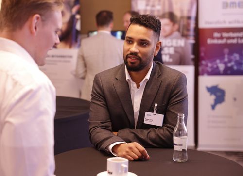 Mann in grauem Anzug mit Namensschild sitzt an einem Tisch und führt ein Gespräch mit einem anderen Mann in weißem Hemd.