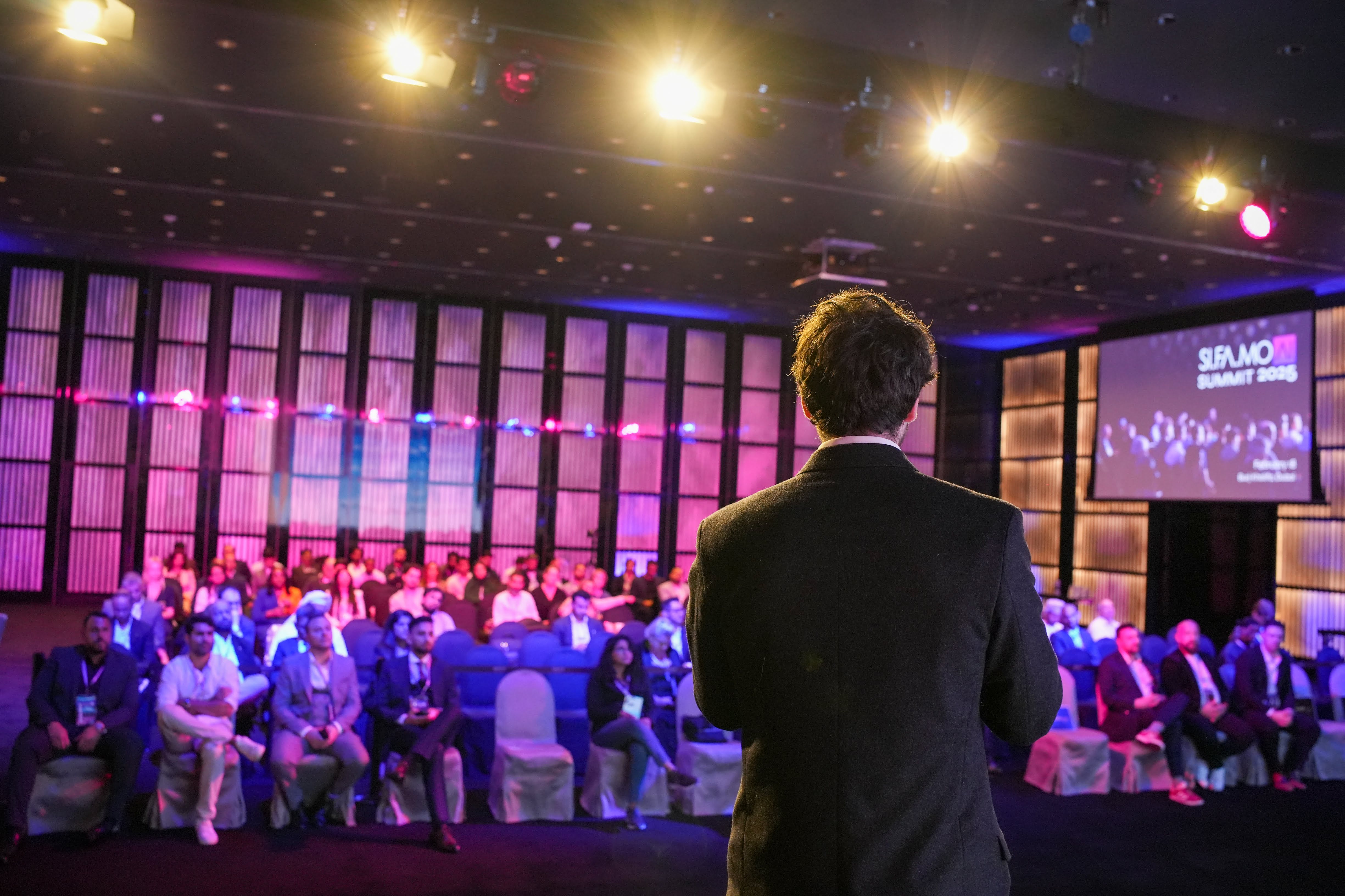 Ein Mann in dunklem Anzug spricht vor einem Publikum bei der SIFAMO Summit 2025 in einem beleuchteten Konferenzraum.