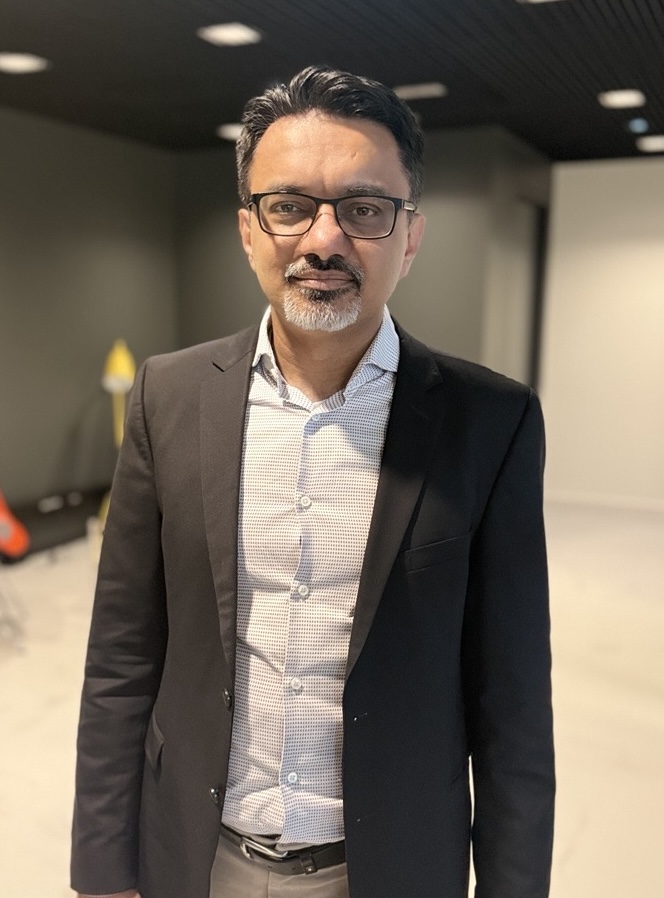 Ein Mann mit Brille, grauem Bart und kurz geschnittenem Haar trägt einen schwarzen Blazer und ein weißes Hemd in einem modernen Innenraum.