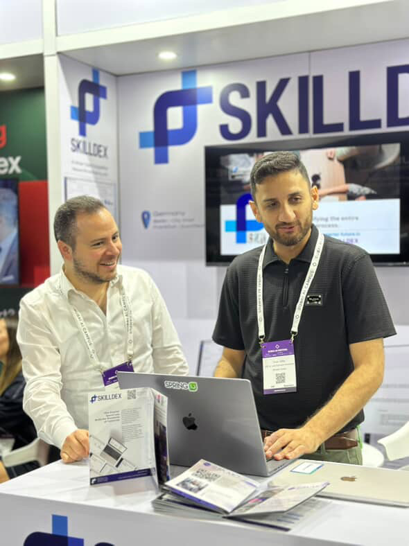 Zwei Männer stehen an einem Messestand mit SKILLDEX-Logo und arbeiten an einem Laptop.