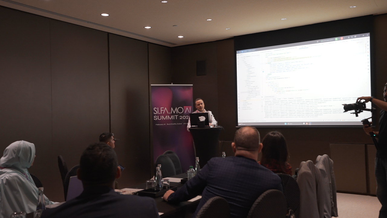 Ein Redner präsentiert bei der SI.FA.MO AI Summit 2023 vor einem großen Bildschirm mit Programmiercode, während Zuhörer in einem Konferenzraum sitzen.