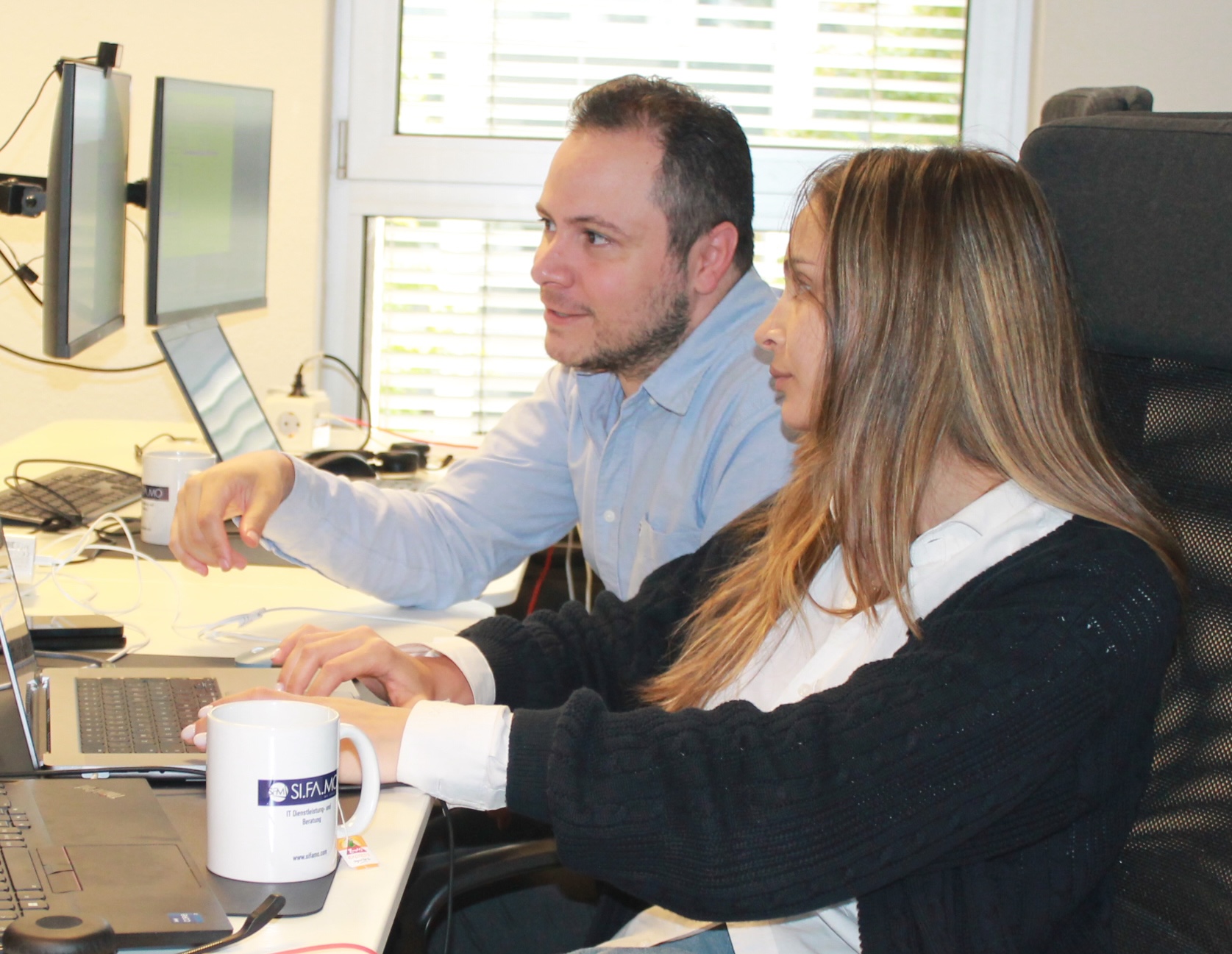 Zwei Personen, die an einem Schreibtisch mit Laptops und Monitoren zusammenarbeiten.