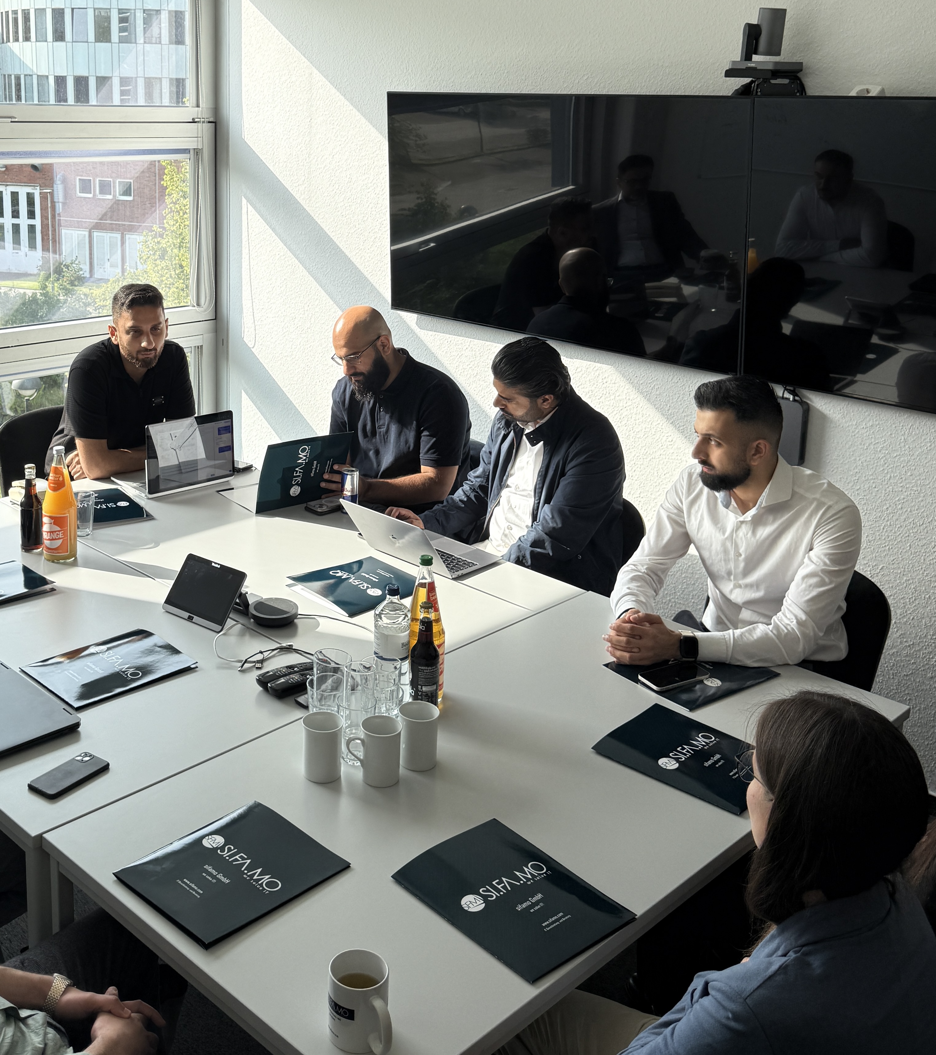 Fünf Personen sitzen um einen Besprechungstisch mit Laptops, Ordnern und Getränken in einem hellen Büro mit großem Fenster.