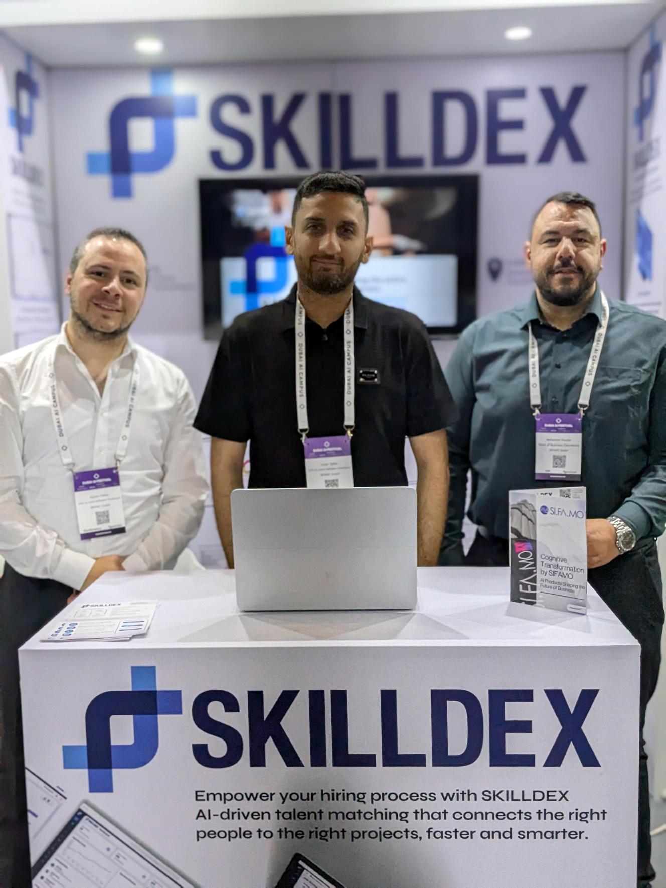 Drei Männer mit Namensschildern stehen hinter einem Messestand mit SKILLDEX-Logo und Laptop.