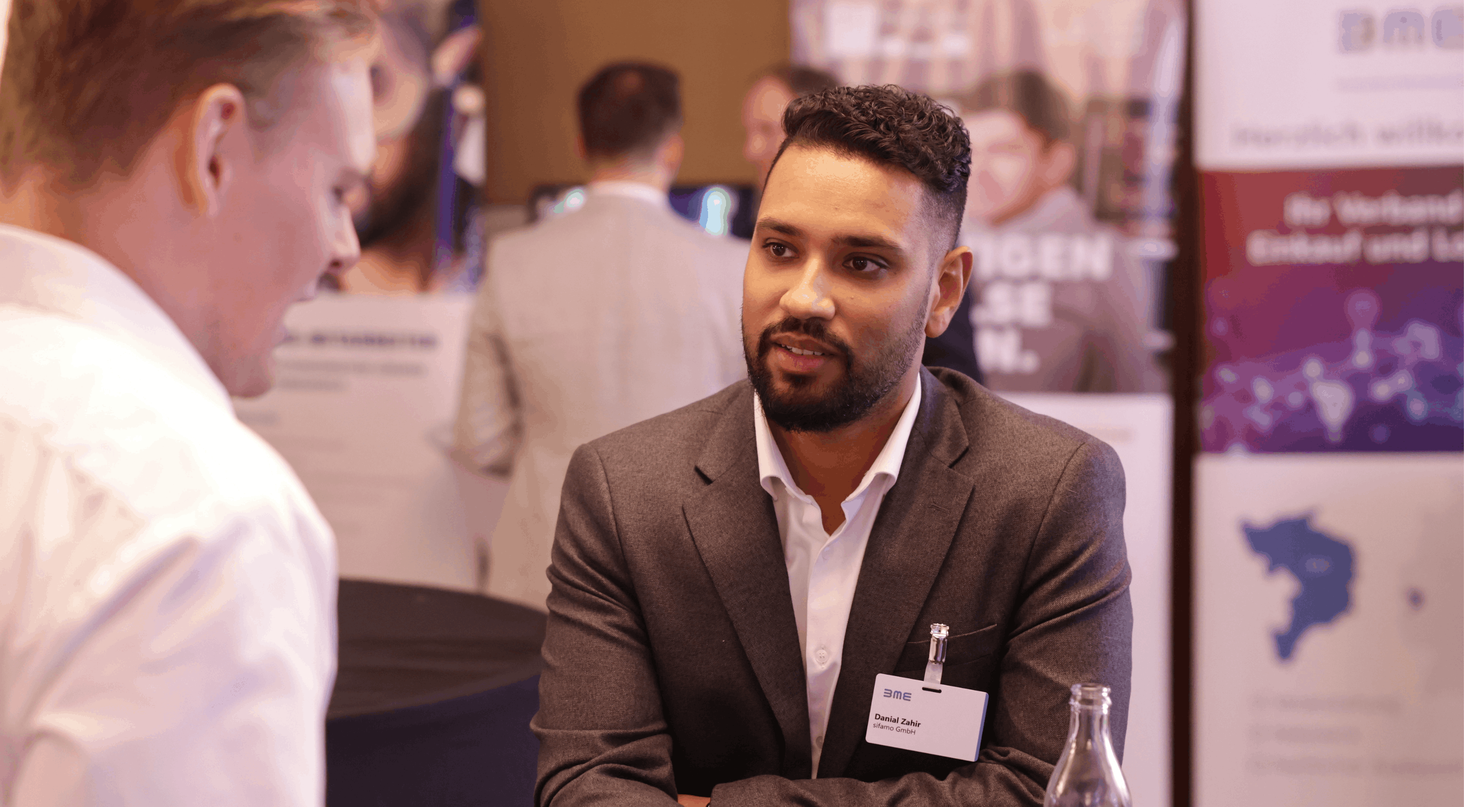 Zwei Männer in formeller Kleidung unterhalten sich bei einer Messe oder Konferenz, einer trägt eine Namensschild.