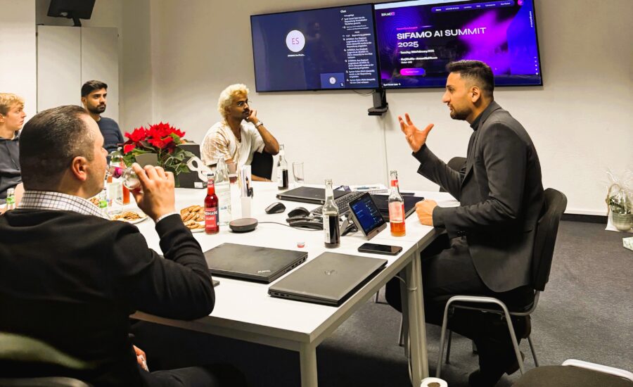 Vier Männer sitzen um einen Tisch, einer spricht und gestikuliert, während die anderen zuhören, im Hintergrund zwei Bildschirme mit einer Präsentation zum SIFAMO AI Summit 2025.